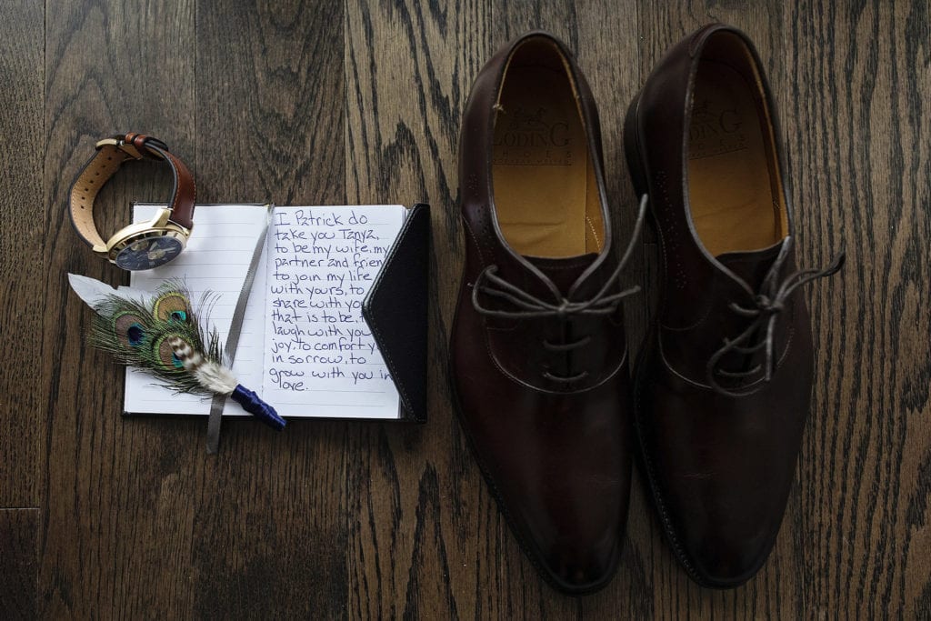 flatlay of groom's shoes, watch, boutonniere and vows notebook
