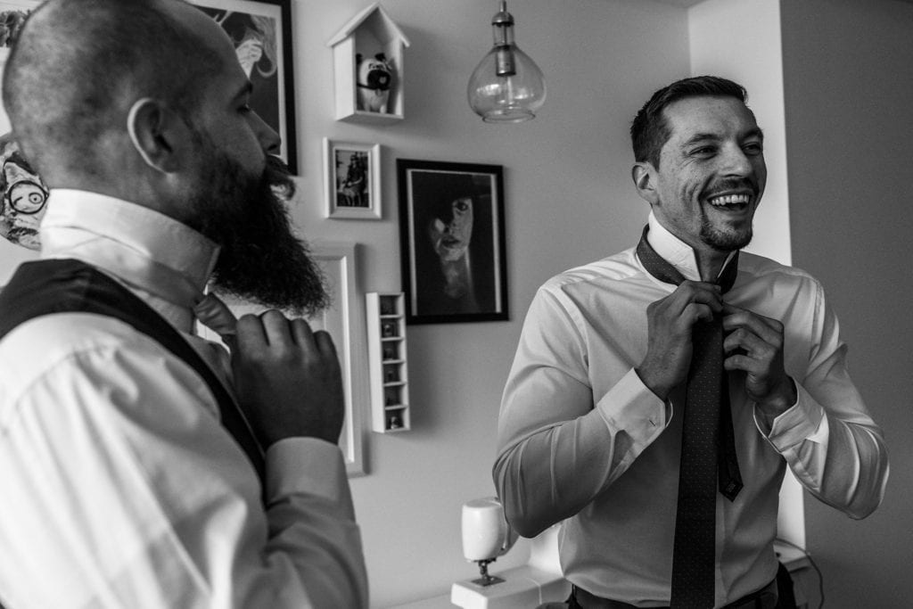 groomsman laughs as he ties tie beside groom during wedding prep