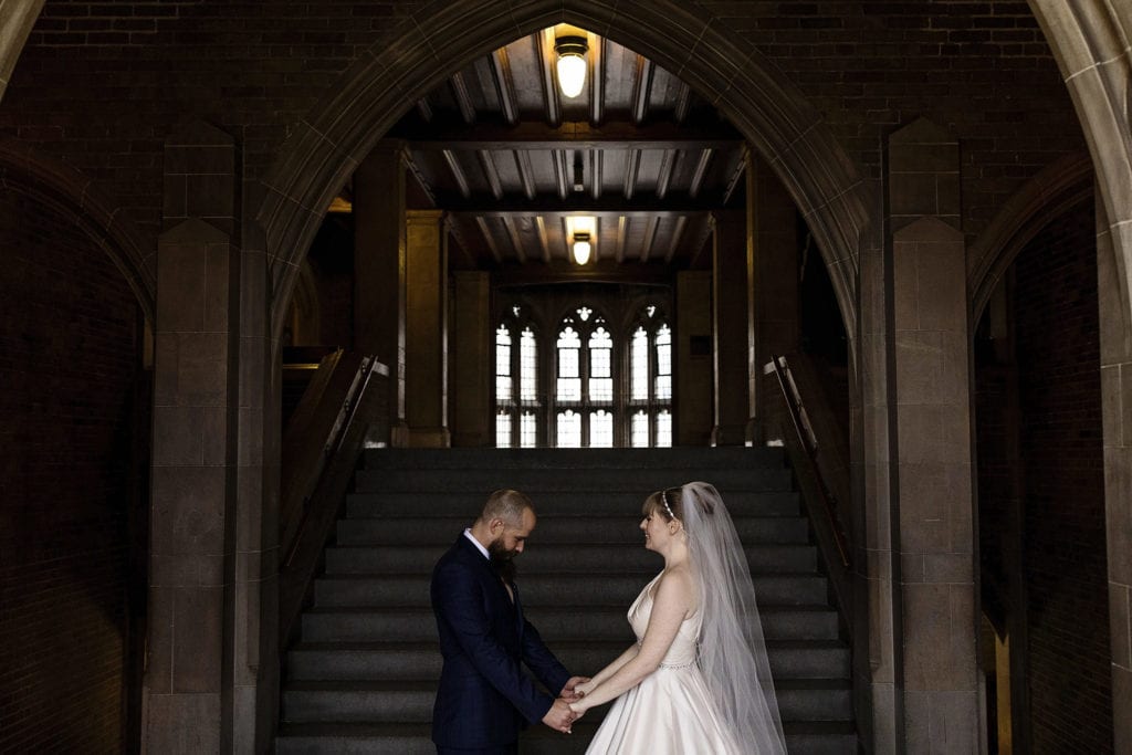 bride and groom hold hands in front of staircase during first look for Hart House Toronto wedding