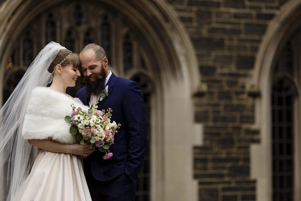 Bride and groom rest heads against each other in ront of sandstone and brick building in Hart House courtyard