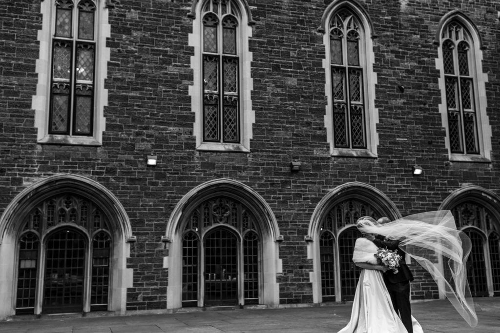 bride and groom stand close together in front of ornate brick building while veil blows across to cover their faces