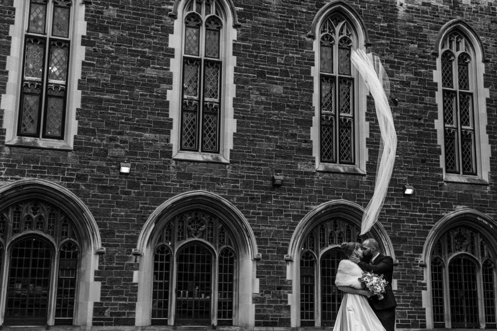 bride and groom stand close together in front of Hart House brick exterior while veil blows vertically
