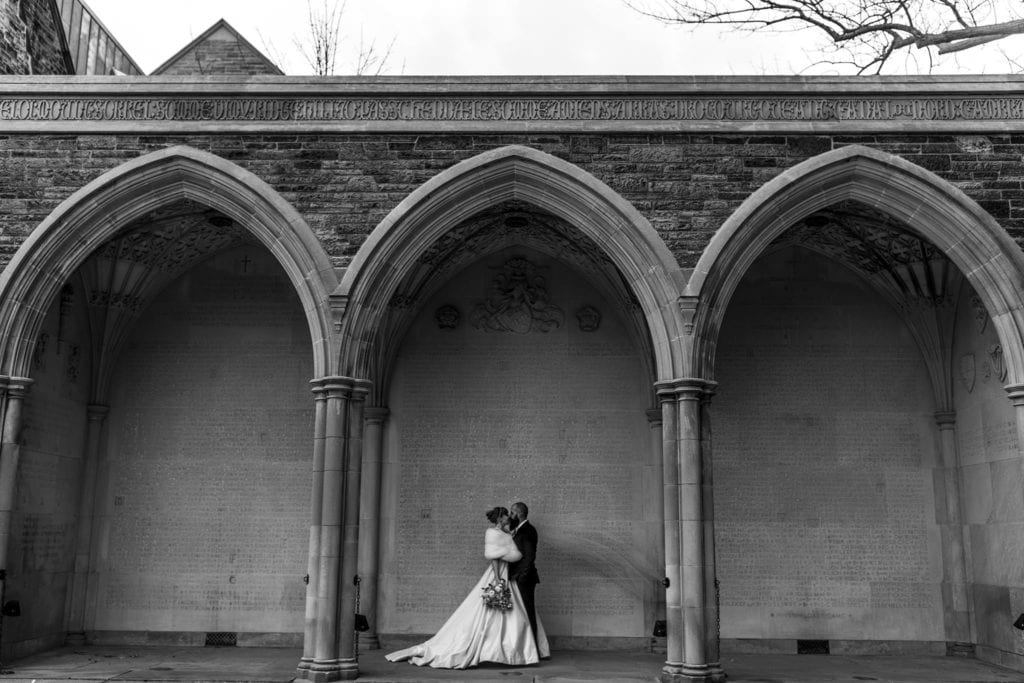 bride and groom stand under arched walkway while veil blows in wind