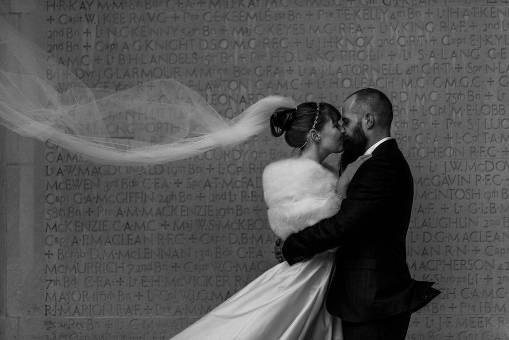 bride and groom kiss in front of text-covered wall while veil ripples in the wind