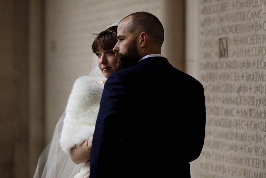 bride in fur stole stands close to groom in front of sandstone wall covered in text
