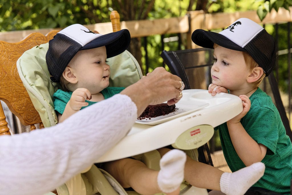 boy in hat rests chin on high chair tray while brother in matching hat eats cake