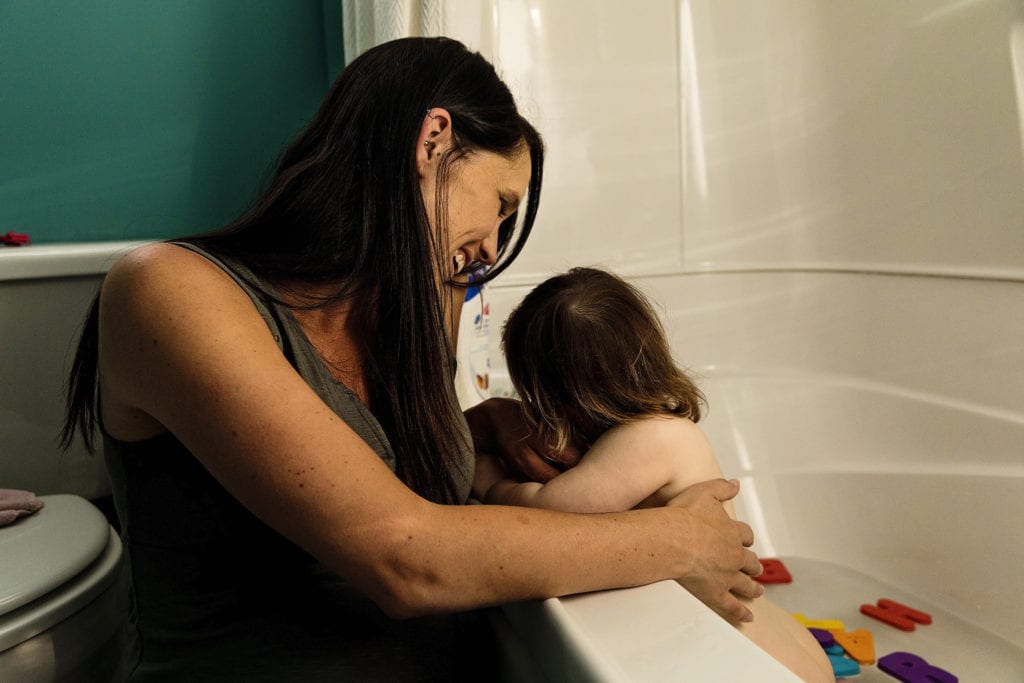 baby in bathtub hugs smiling Mom's hand