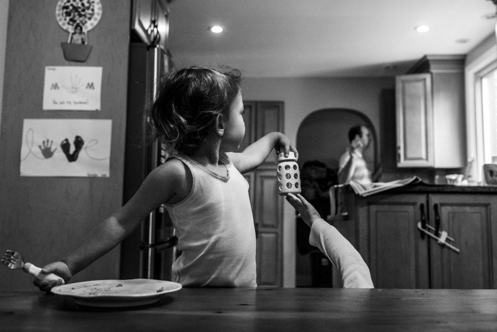 girl in white tank top reaches cup out while arm extends from below table to touch cup