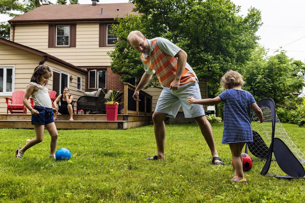 dad plays soccer with two girls while mom sits on step and watches