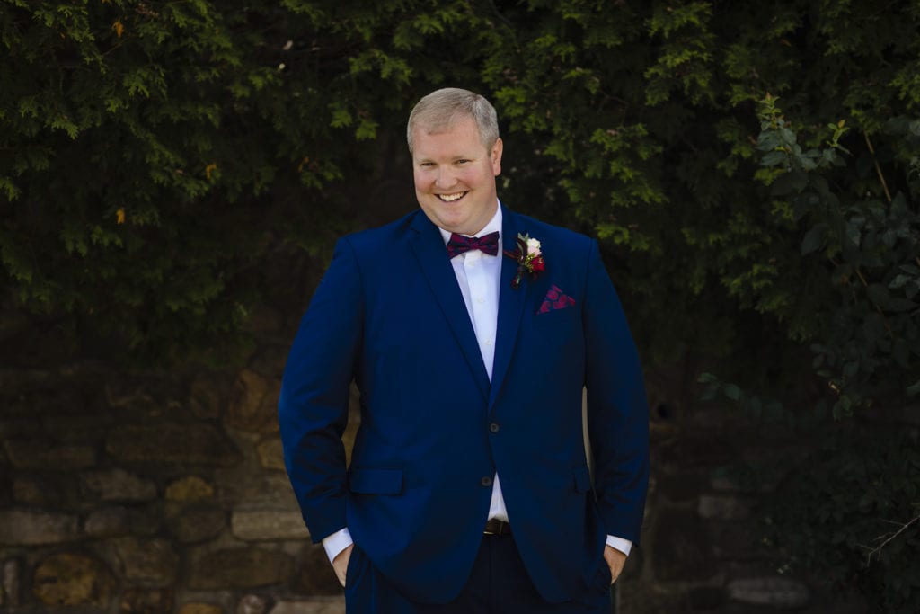 groom in royal blue with bow tie stands with hands in pockets in front of cedars