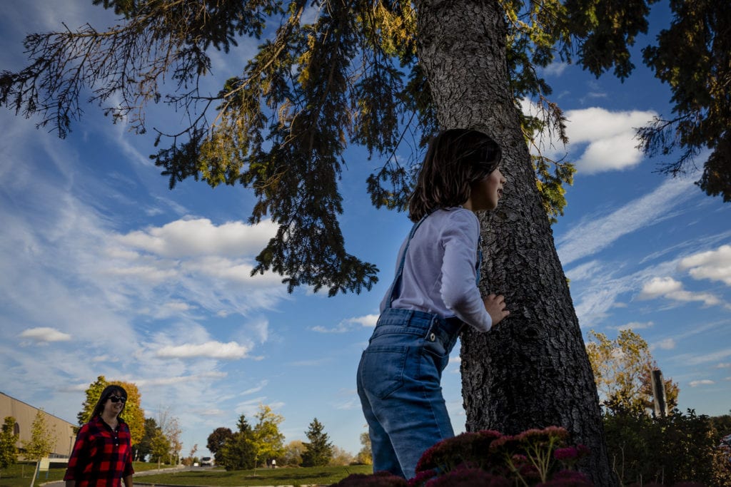 girl in dungarees peeks out from behind pine tree stump