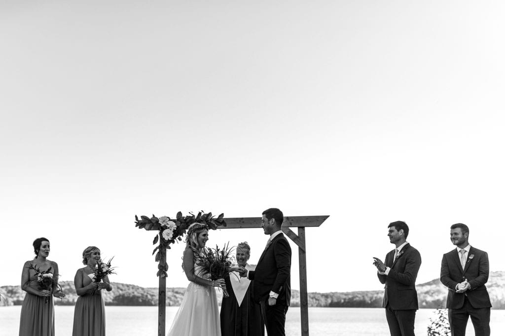 bride and groom laugh while holding hands in front of officiant and wedding party during wedding ceremony at the lake