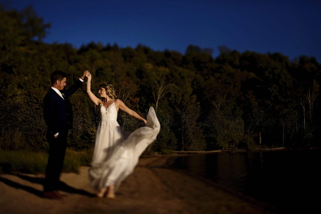 groom holds bride's hand as she twirls and dress flies up on the sand next to lake