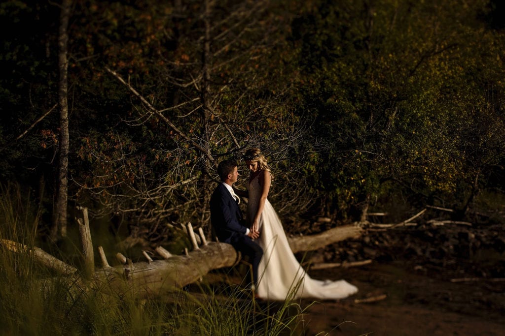 groom sitting on log by lake holds hands with bride who stands looking down at him