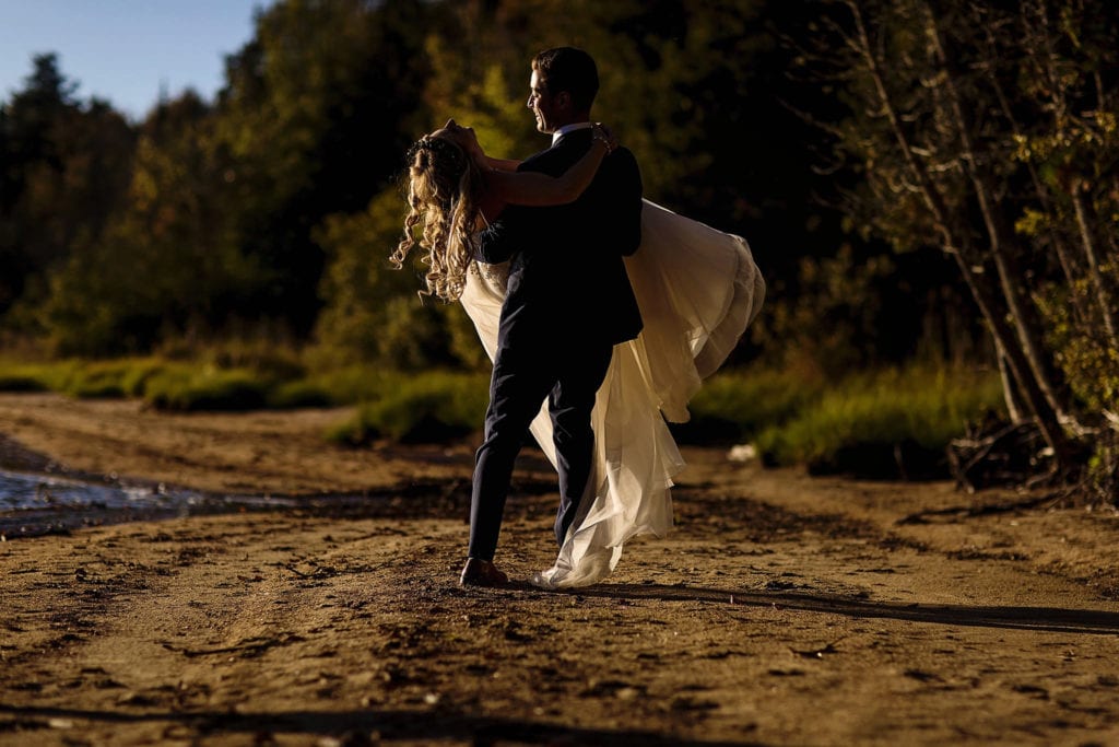 groom on lakefront beach carries bride wearing flower crown who throws her head back