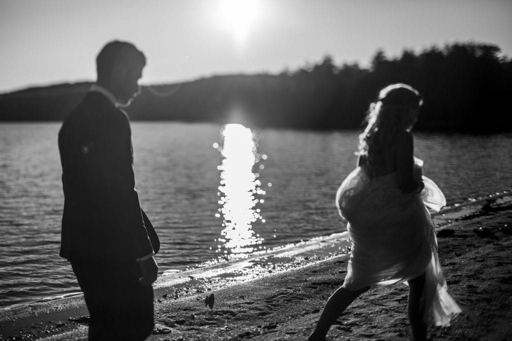 bride and groom walk along lake with sun shining while bride carries tulle skirt high in the air