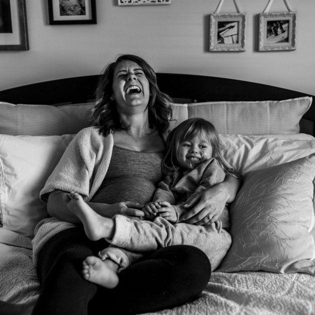 pregnant woman sits on bed and laughs while holding daughter
