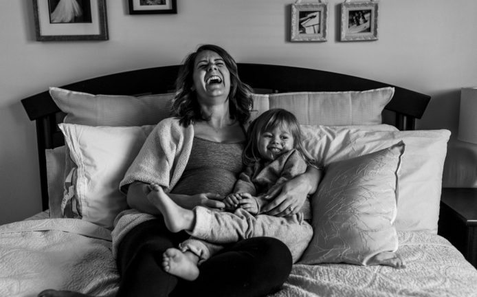 pregnant woman sits on bed and laughs while holding daughter