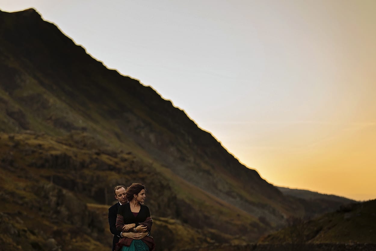 couple stand in valley at dusk looking opposite ways during snowdonia portrait session