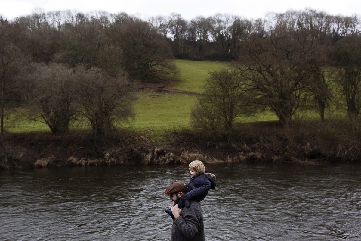 father with flatcap walks with son on shoulders framed in river during documentary session with ontario family photographer