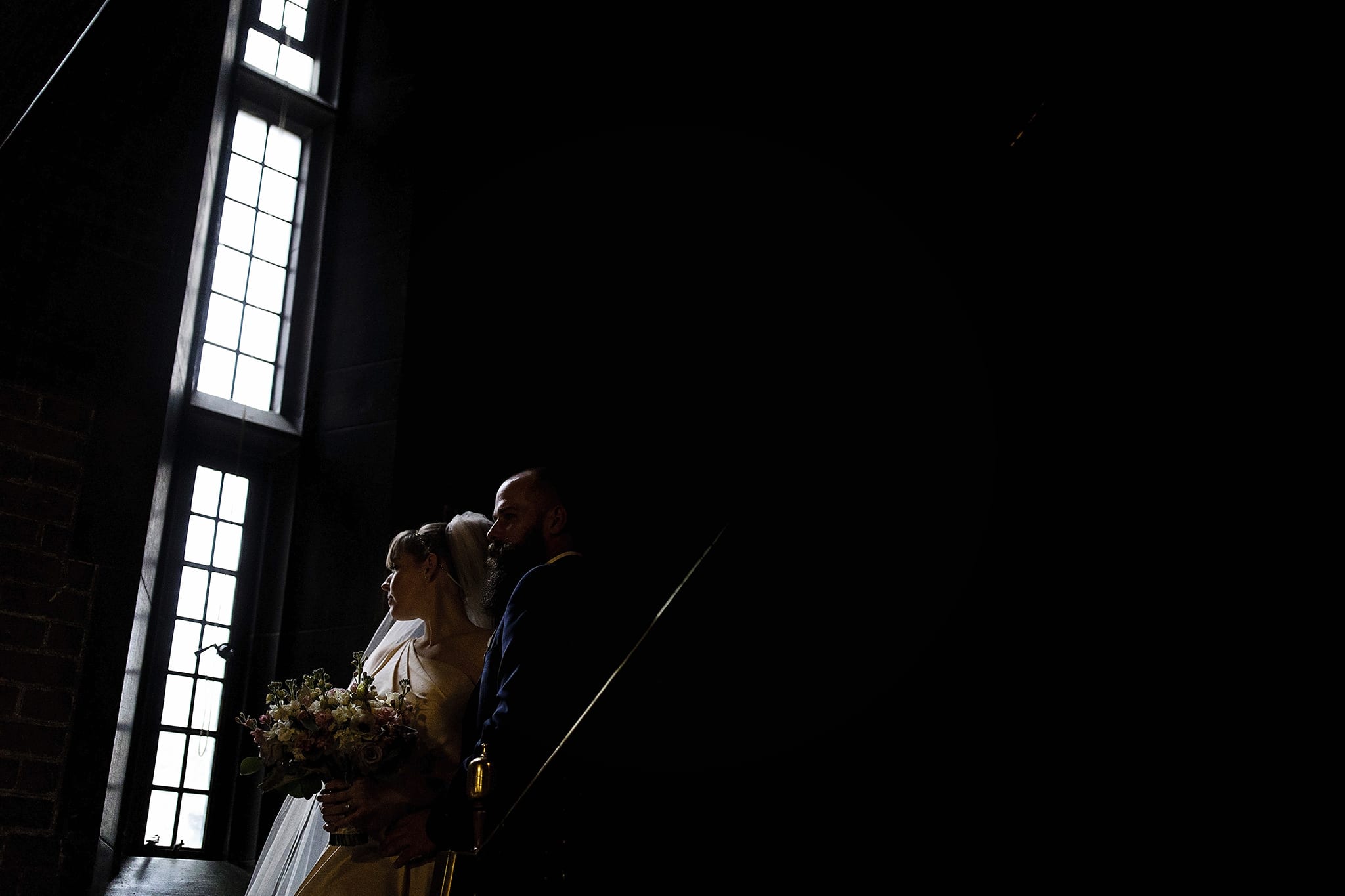 bride and groom stand on stairs lit by dramatic window light