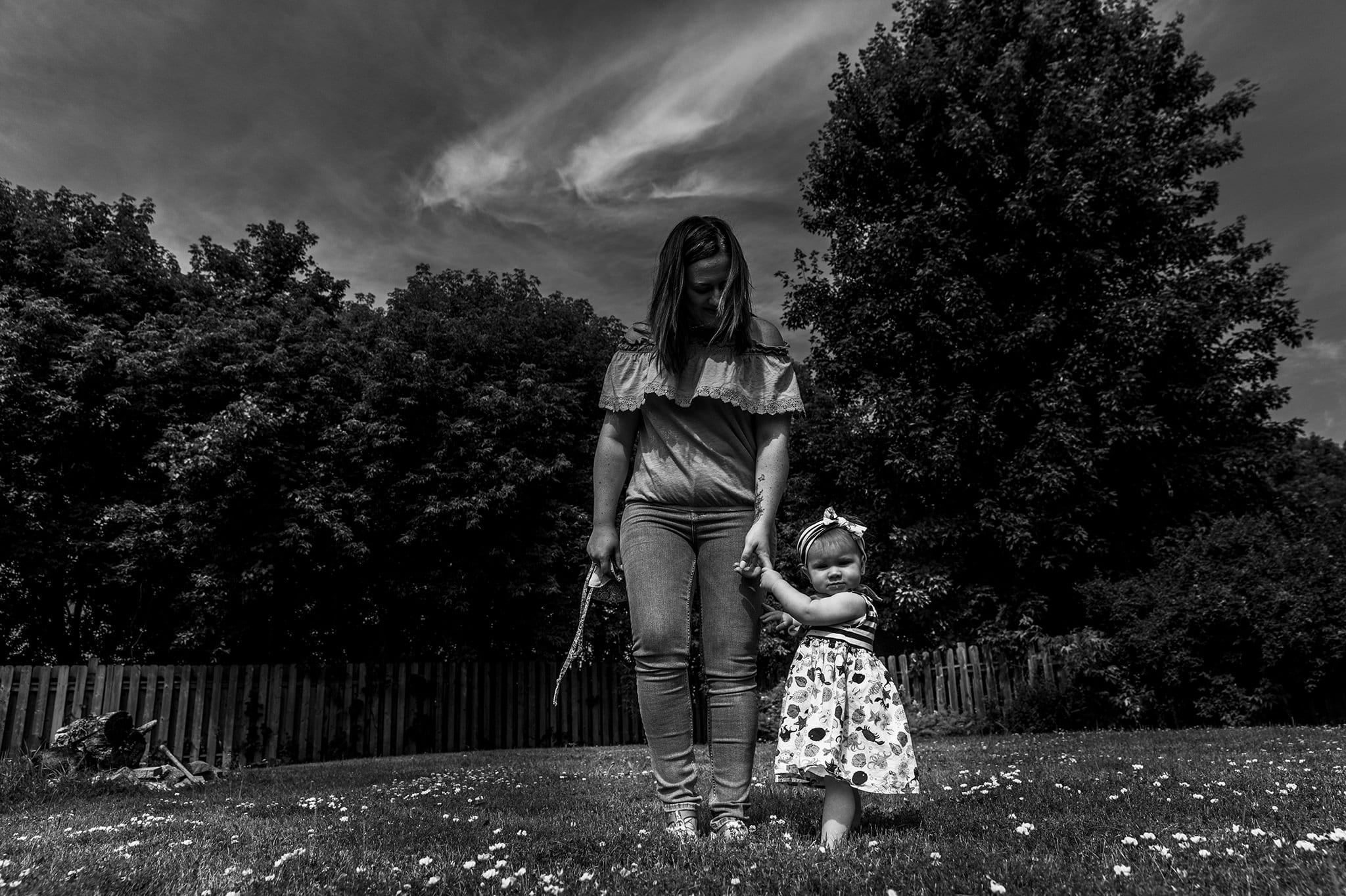 little girl in dress stands in yard holding mother's hand