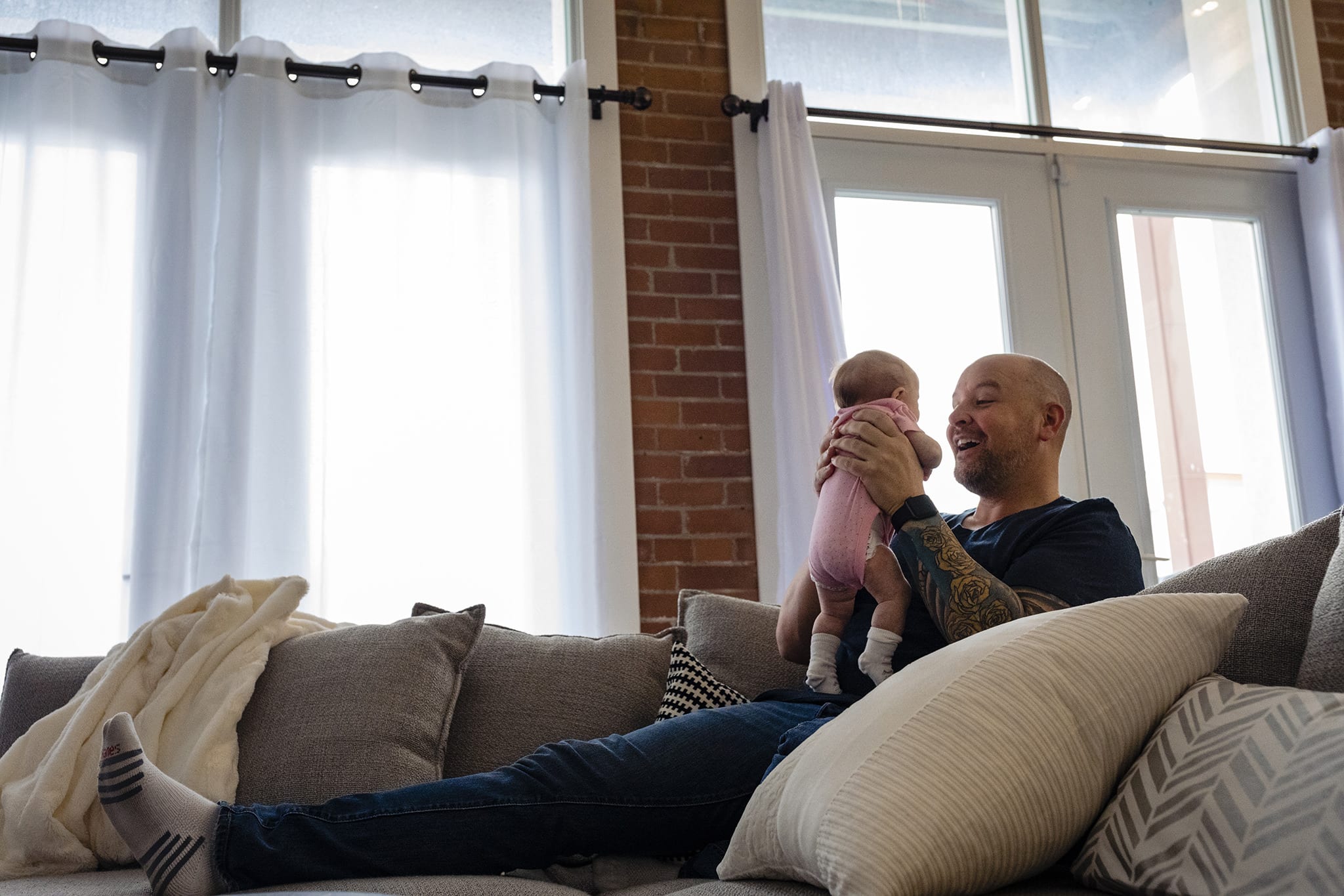 Man sits on couch in front of large windows holding baby in pink onesie during Cotton Mill Loft At-Home Newborn Session