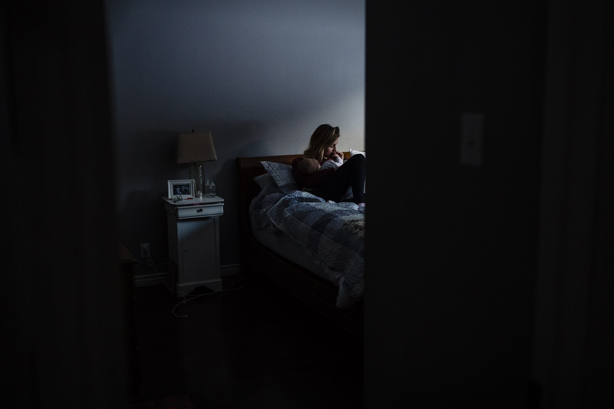 view from doorway of mother in blue light of afternoon breastfeeds baby on bed