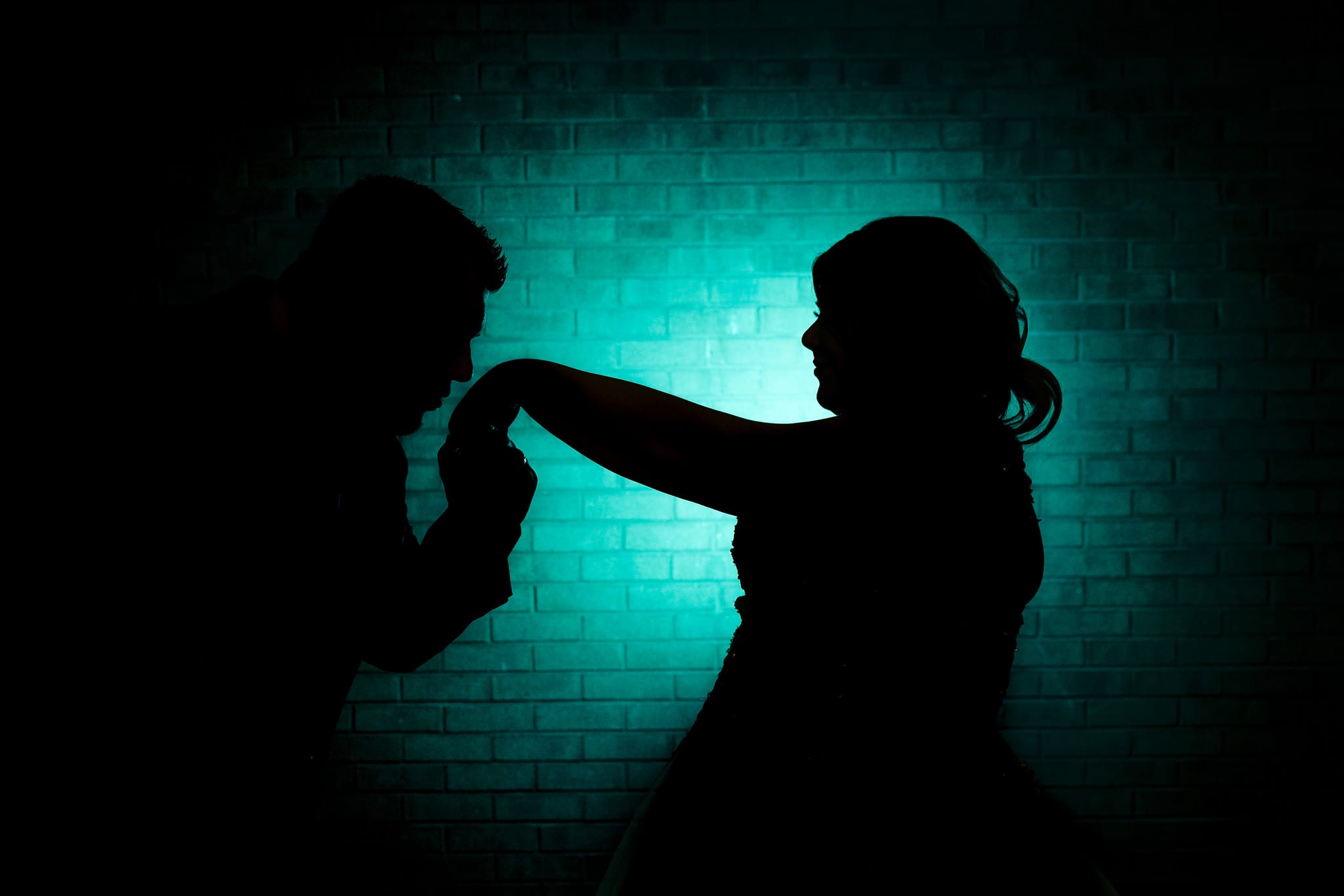 groom kisses bride's hand in silhouette in front of turquoise brick wall