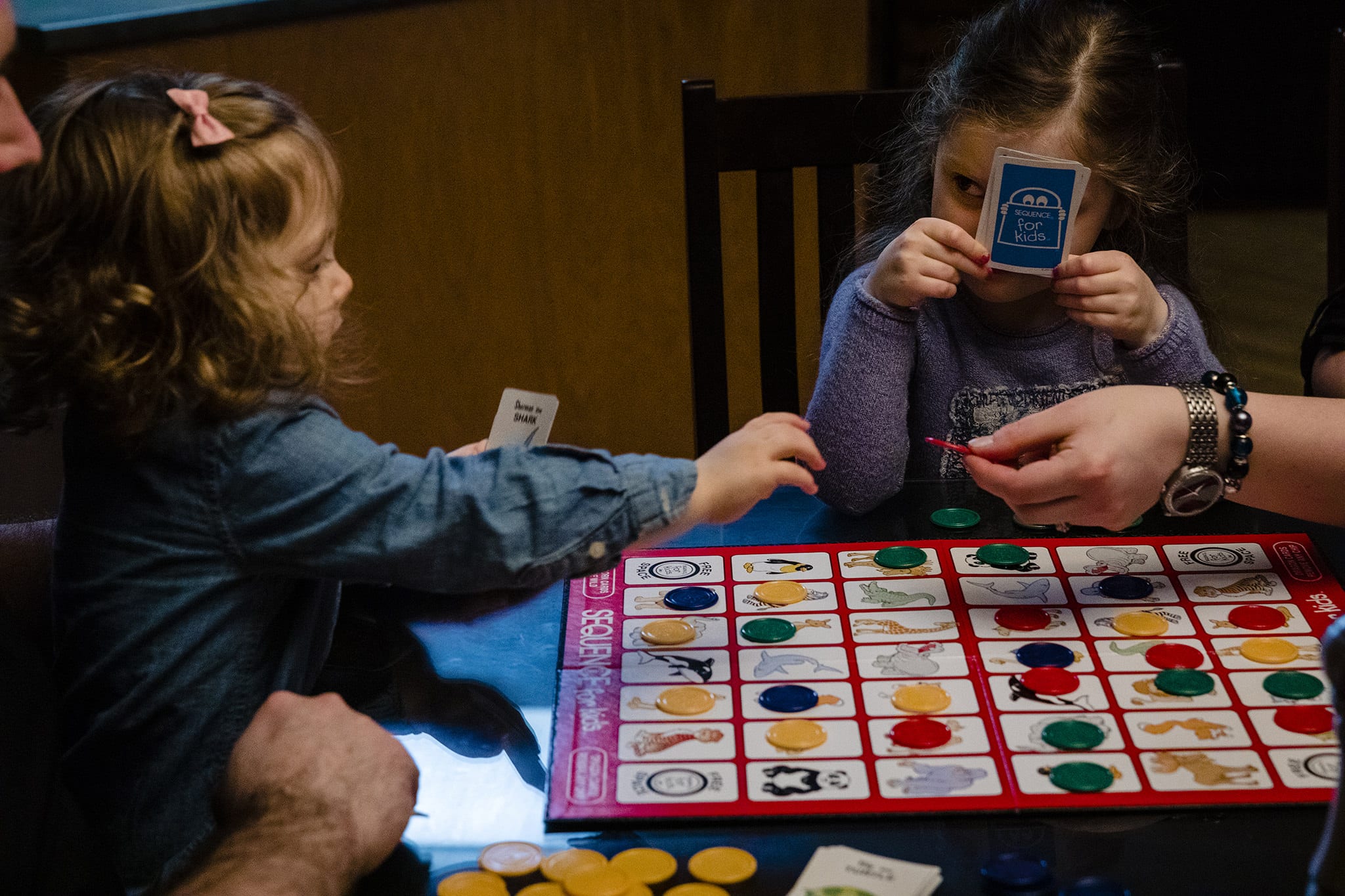 girl peers from behind playing card while toddler reaches for game chip