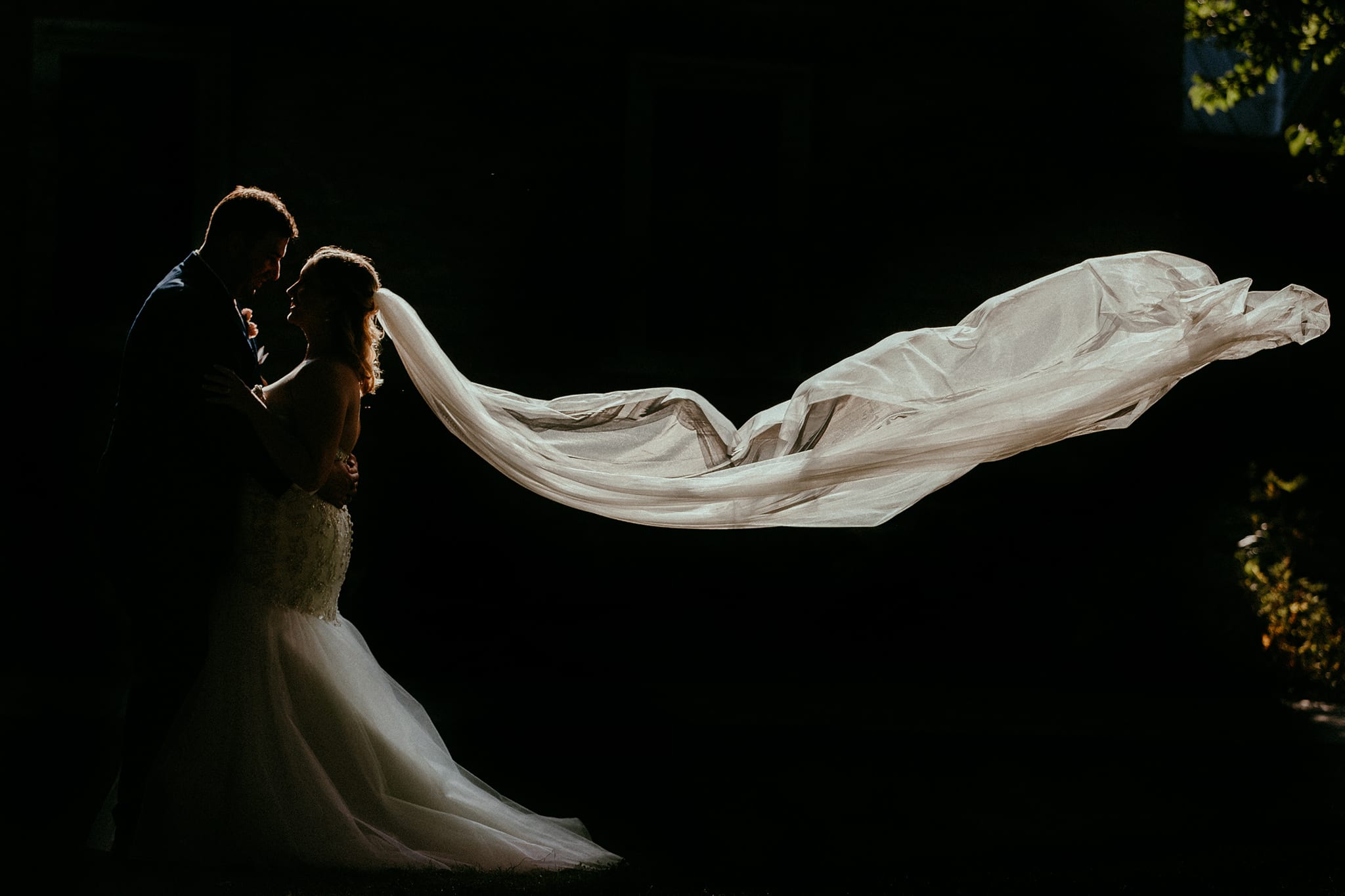 bride and groom in backlit silhouette against black background with veil flying