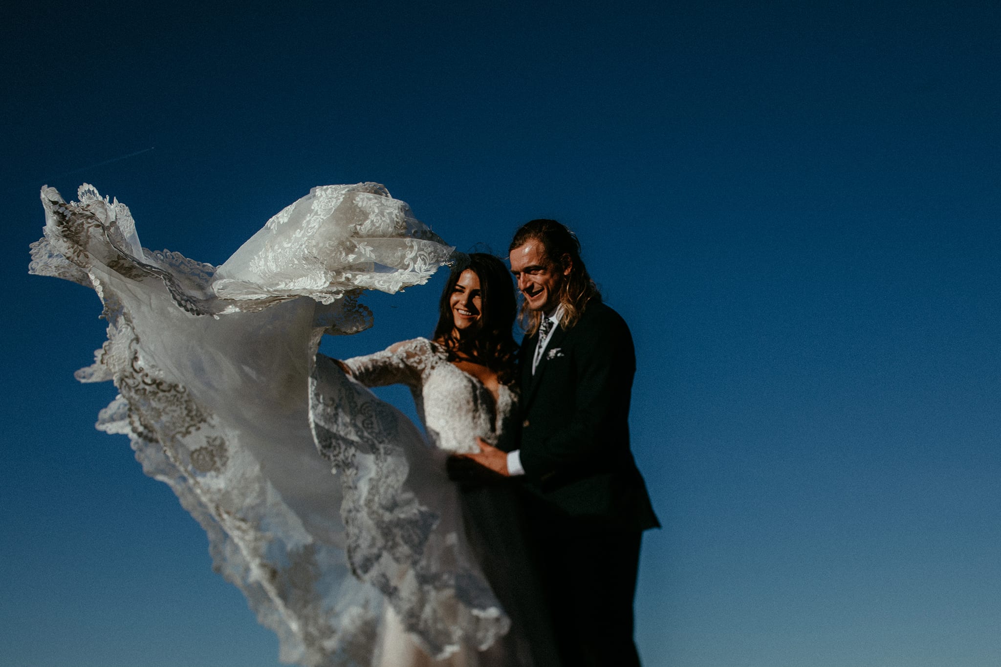 Bride and groom smile against blue sky while lace train flies up in the air