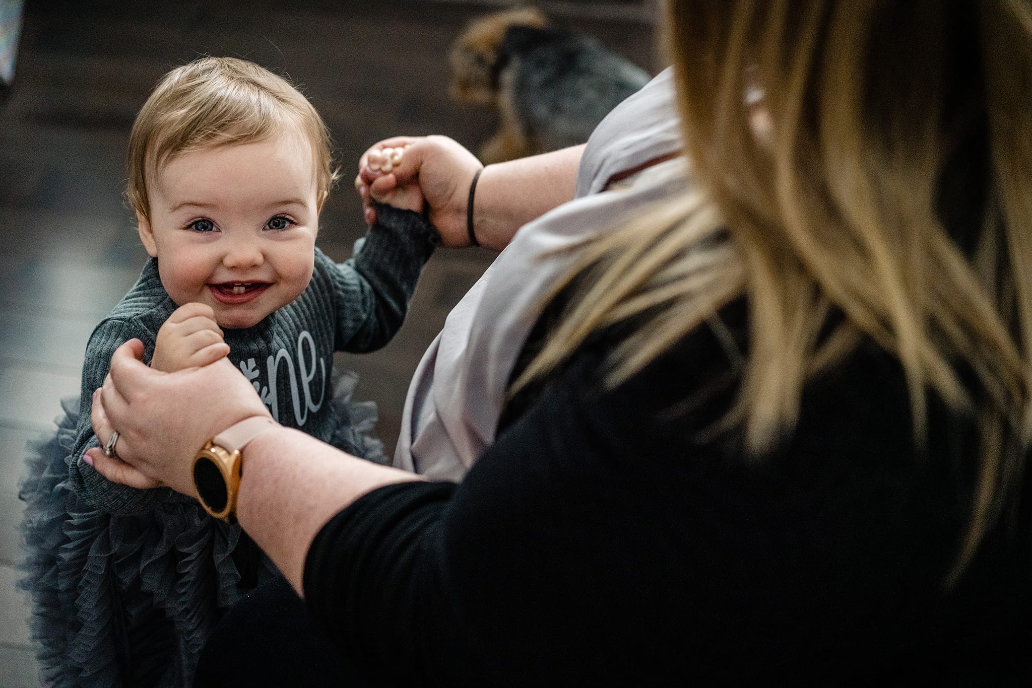 smiling toddler looks at camera while dressed in wintry first birthday clothes and holding mom's hands