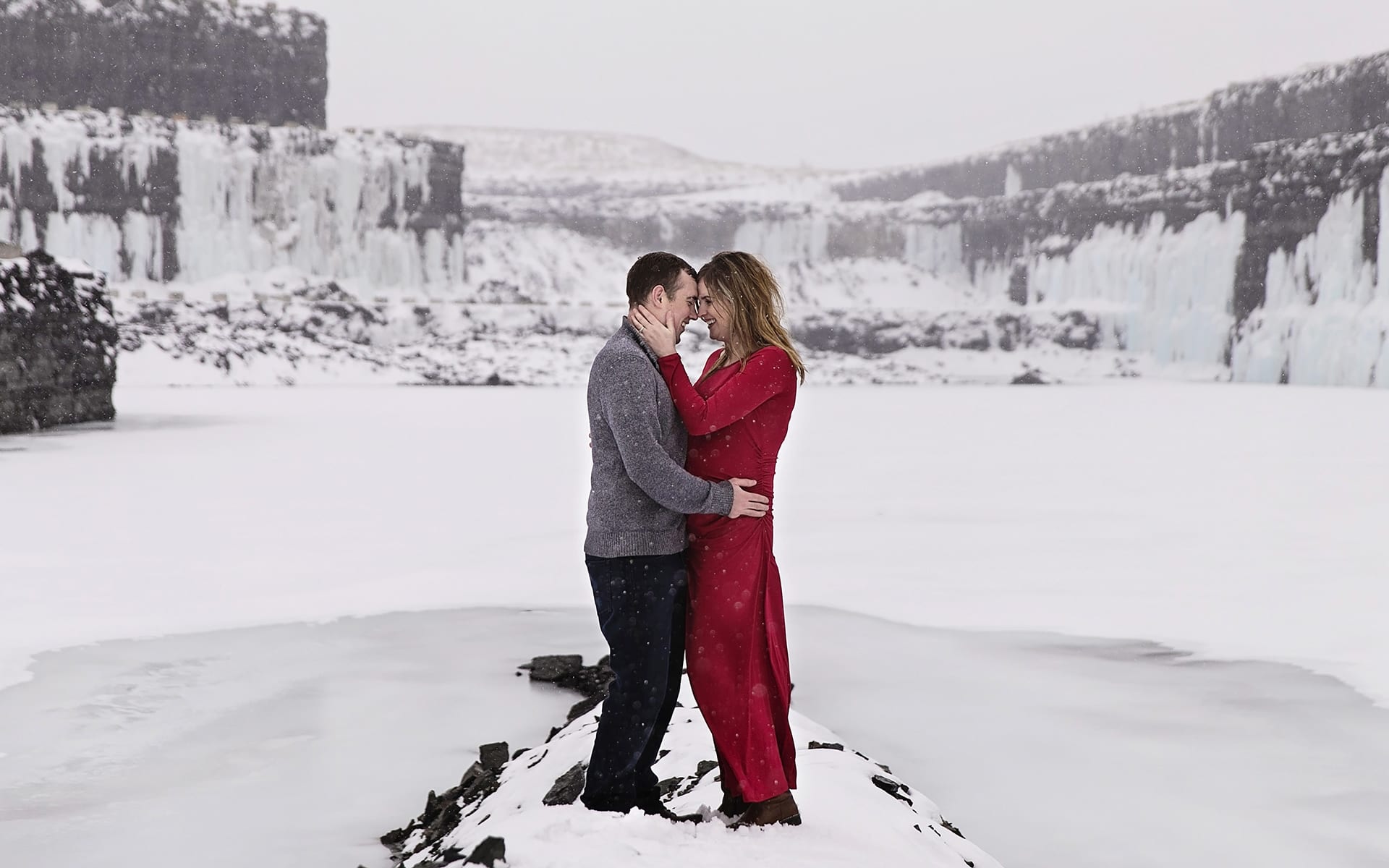 pregnant woman in red maternity dress holds husband close in icy quarry