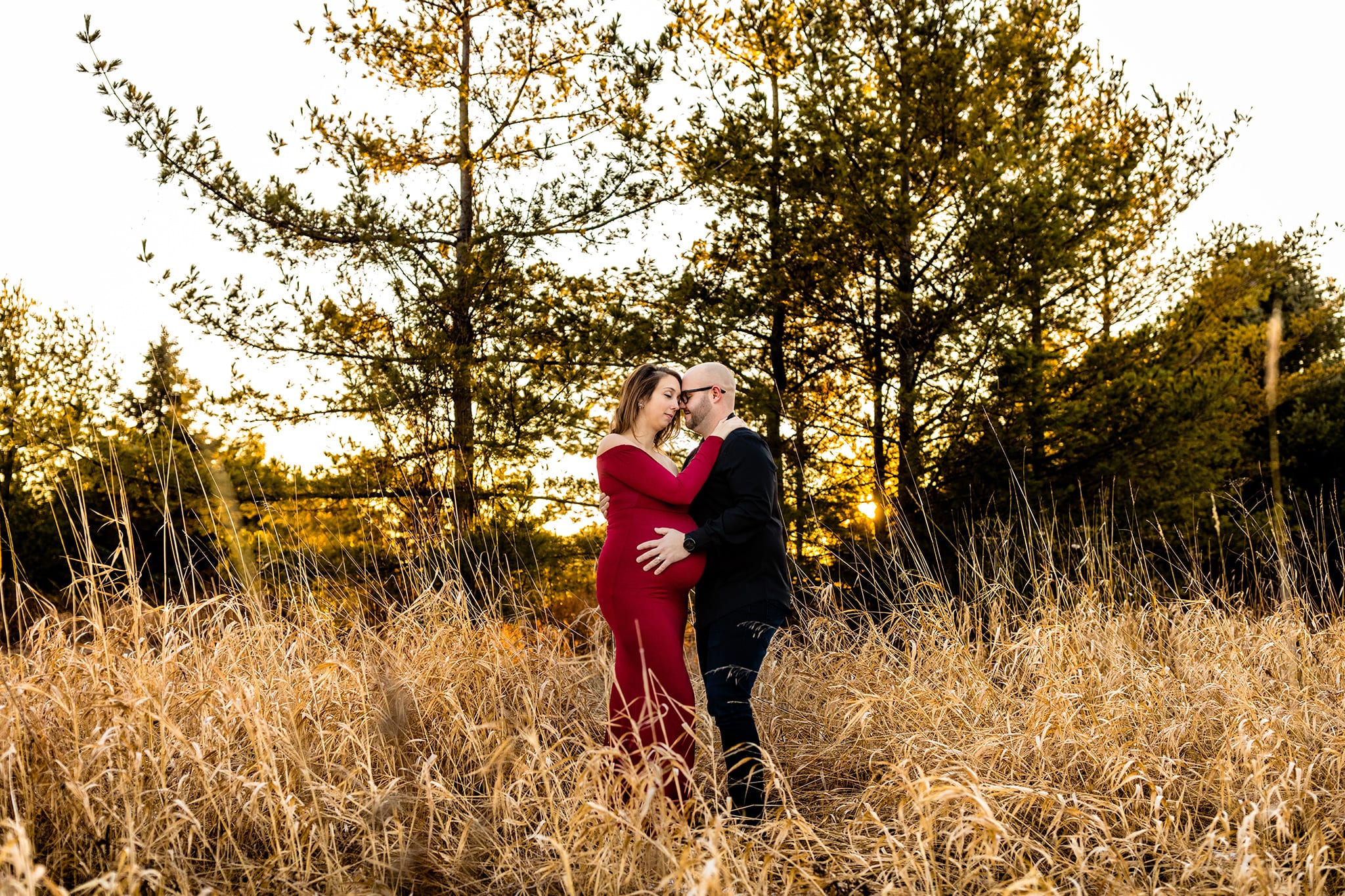 pregnant woman in red dress stands with husband in black shirt in tall grass near pine trees