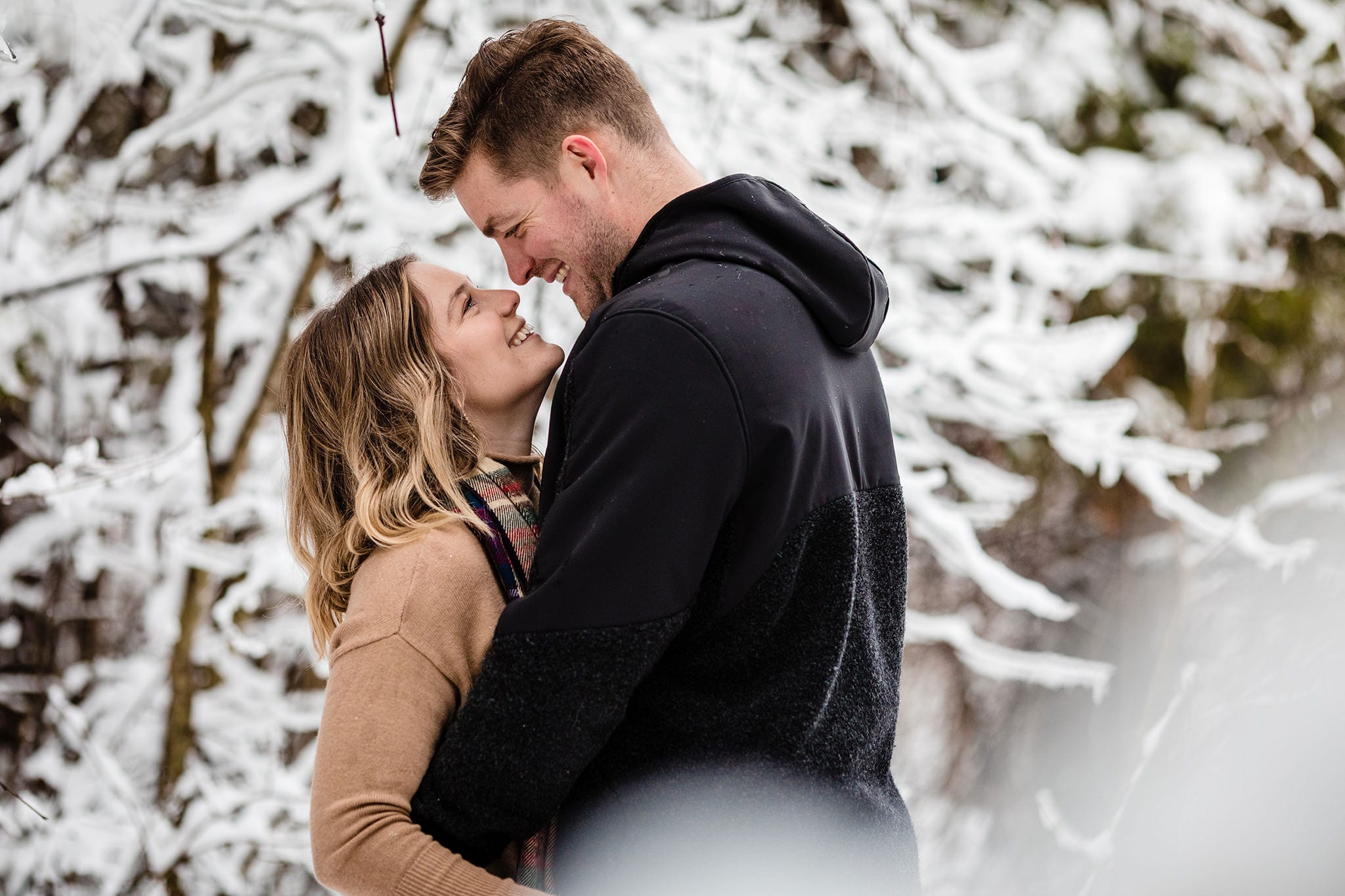 man and woman stand close together while making eye contact in front of snowy trees