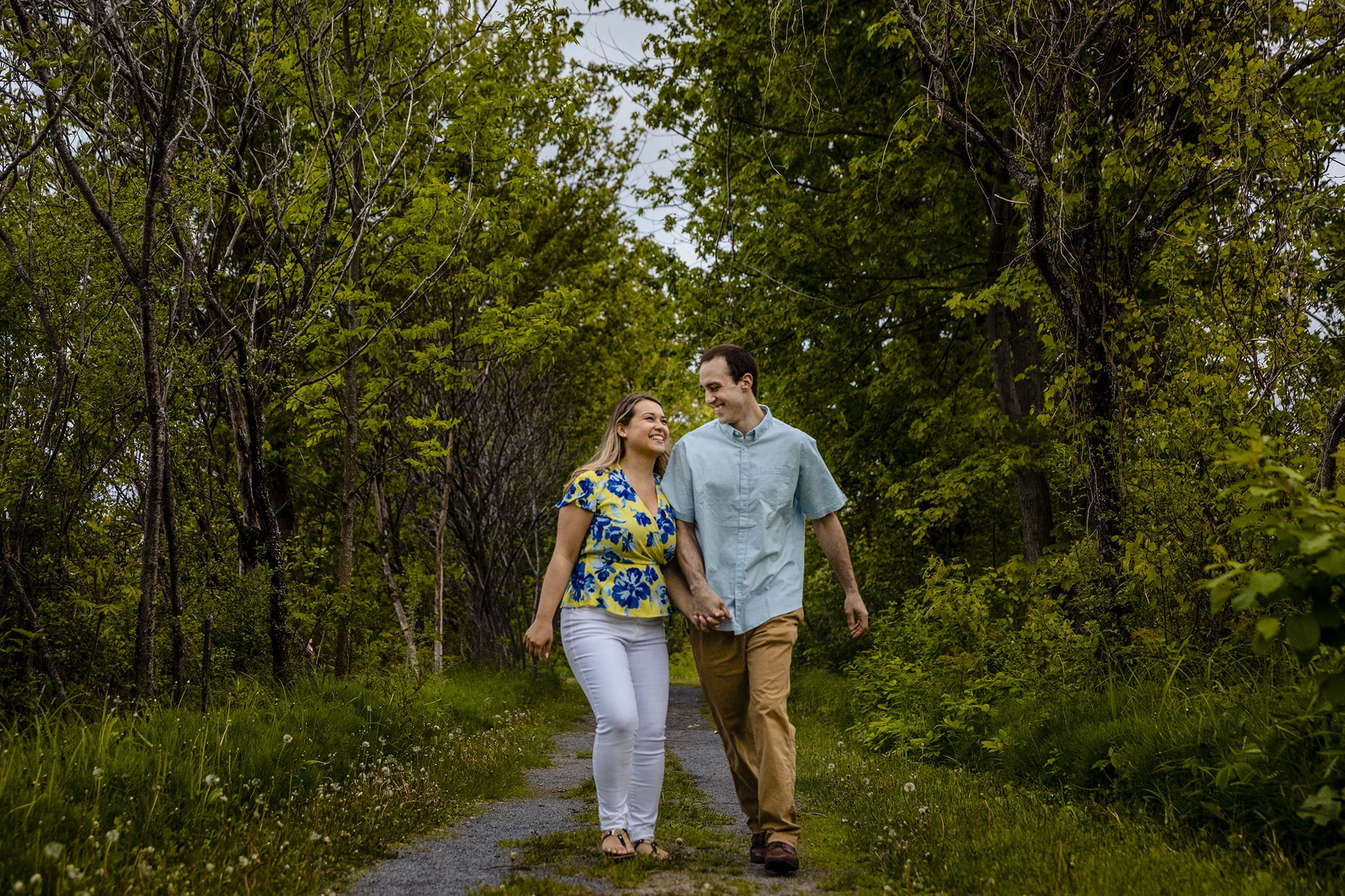 man and woman laugh while walking and holding hands on forest path