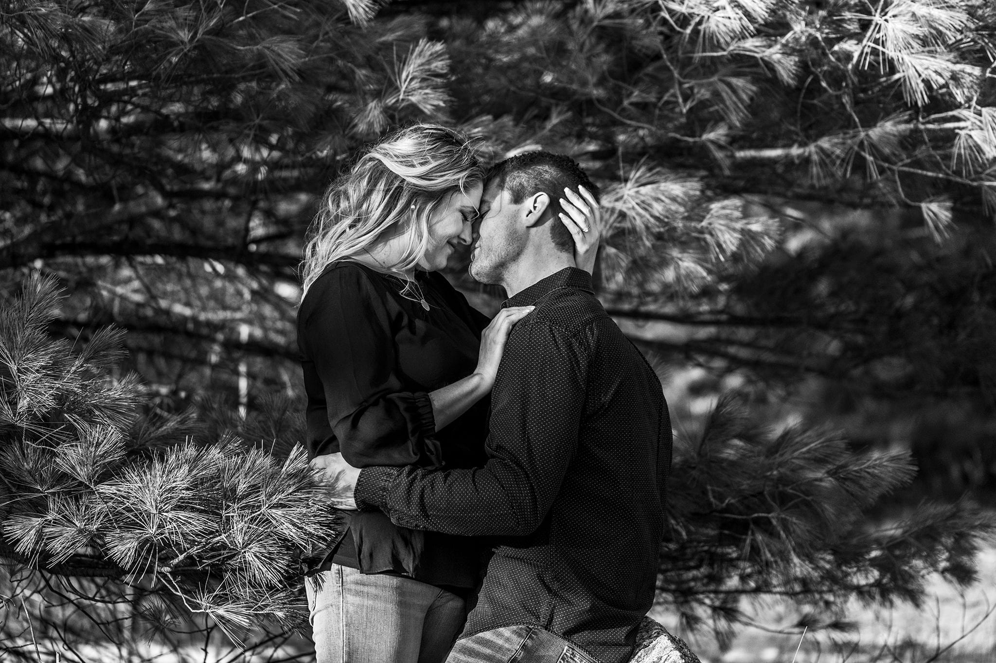 woman leans in to kiss fiancé while surrounded by pine branches
