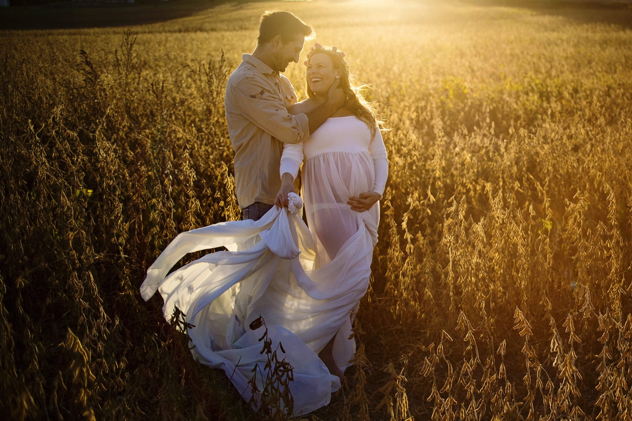 pregnant woman in billowing white maternity dress in soy field looks at man touching her neck