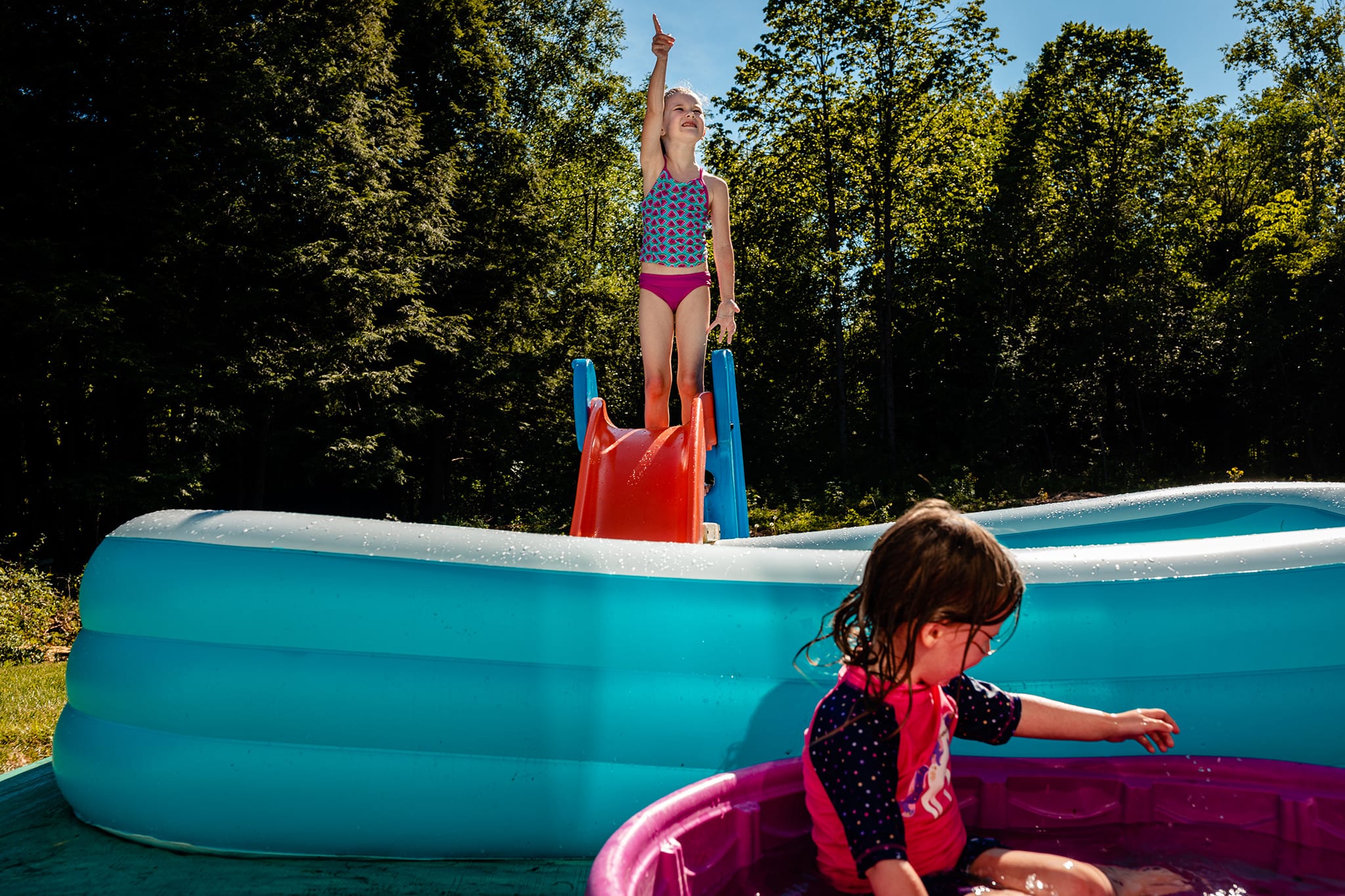 girl in pink and blue bathing suit points at sky while standing on plastic slide and sister in fuschia kiddie pool plays with water