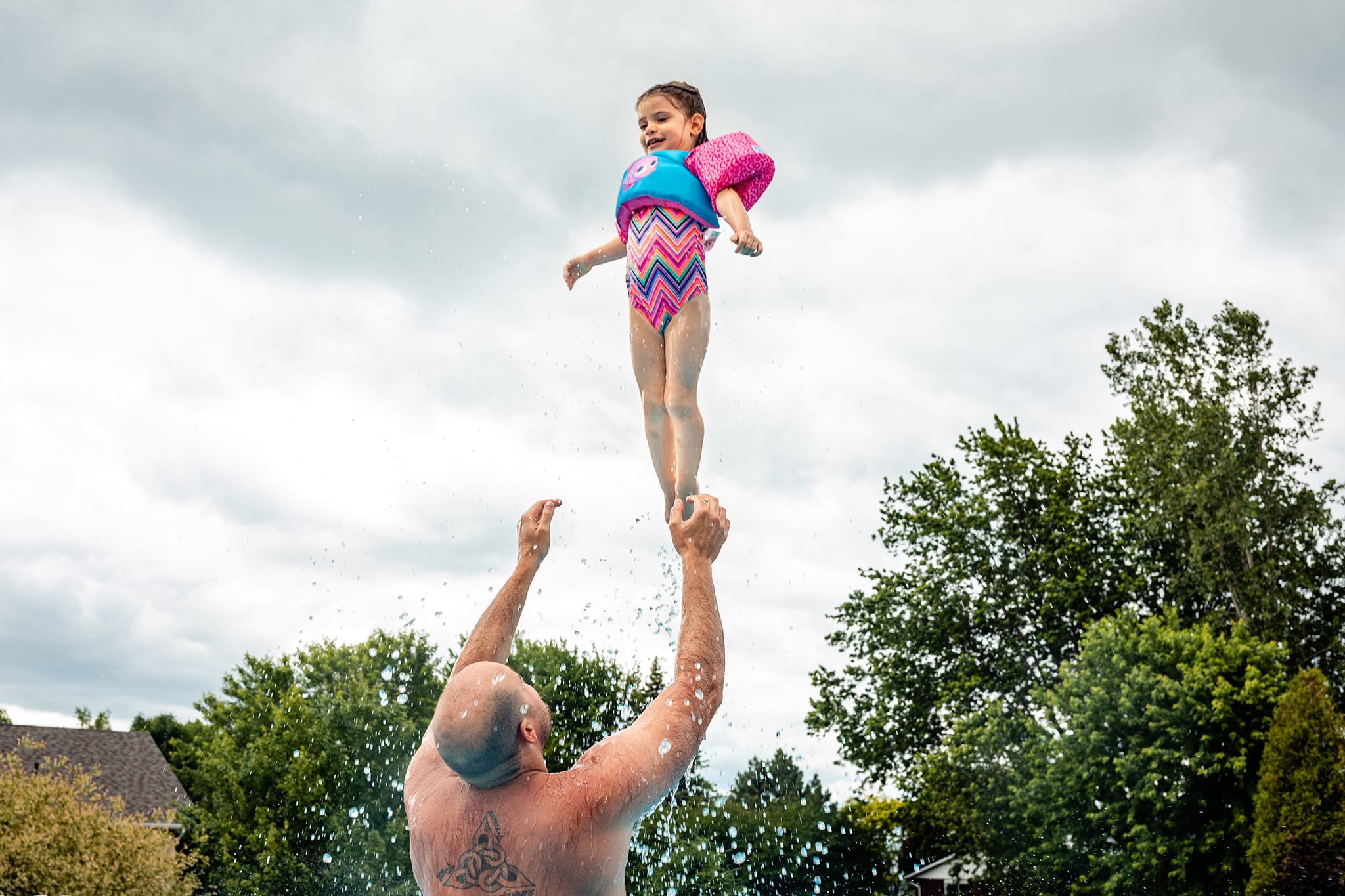 dad with Celtic back tattoo throws daughter in pink puddle jumper and chevron bathing suit high into cloudy sky