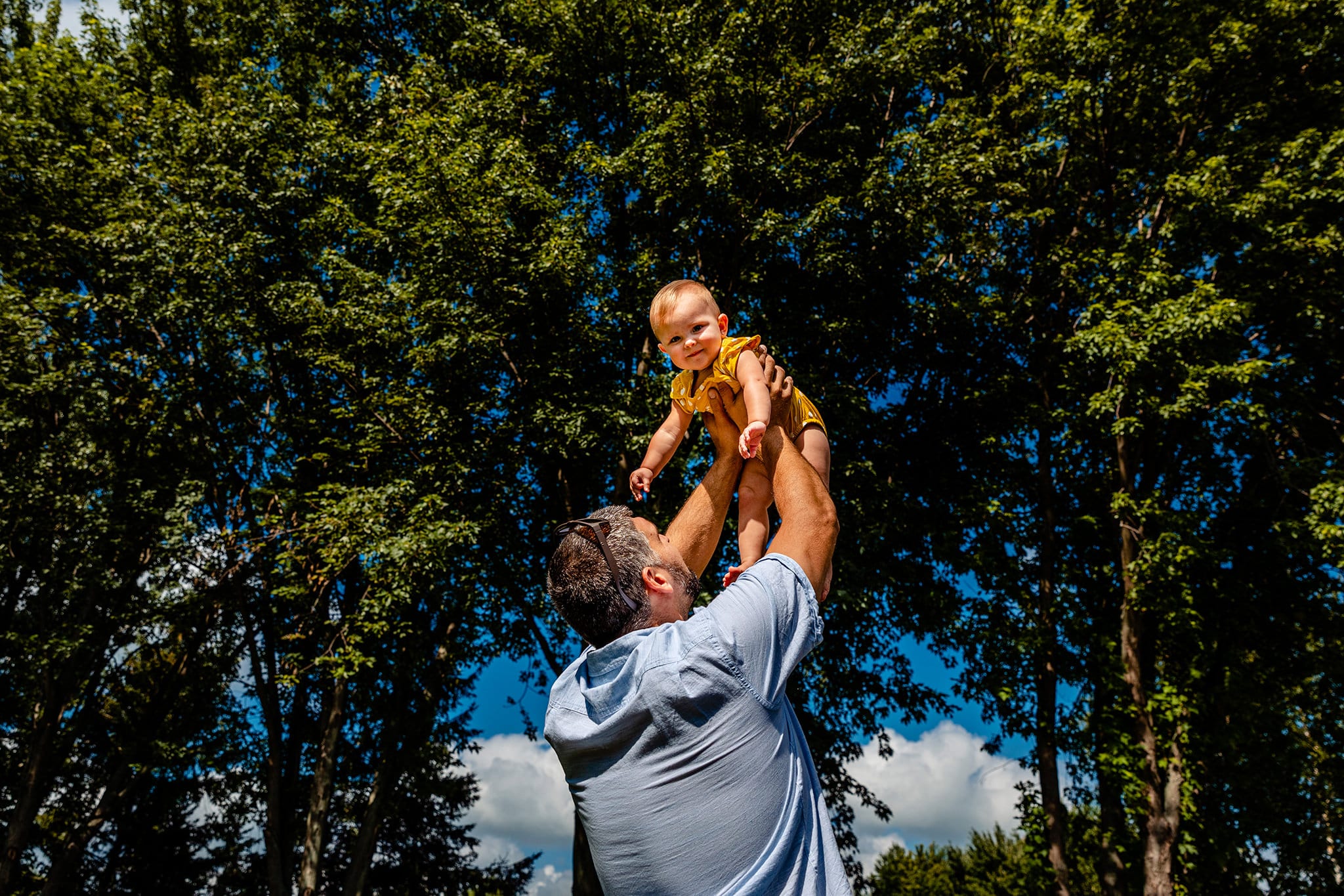 father in blue shirt holds toddler daughter in mustard and white polka dotted romper up in air during cornwall-first-birthday-photos