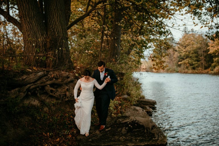 groom helps bride navigate rocks along bank of the Raisin River