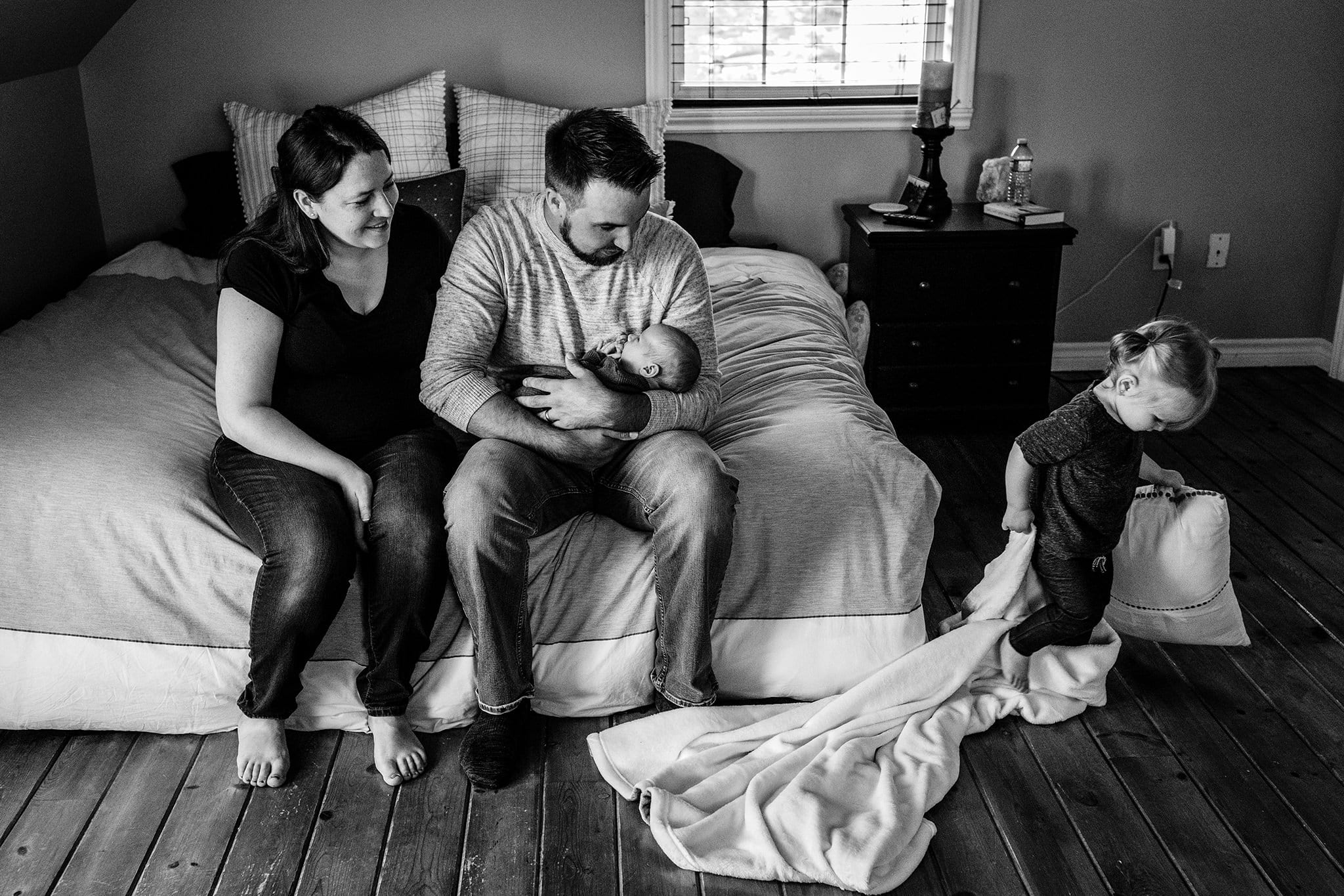 toddler carries blanket and pillow past mom and dad holding baby on bed