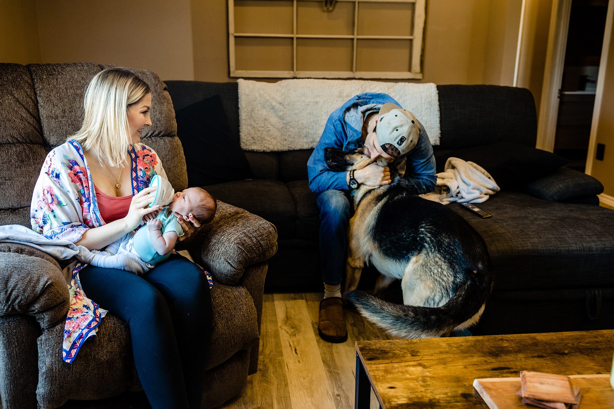 dad in ballcap plays with dog while mom feeds newborn with bottle during at-home Cornwall newborn photos