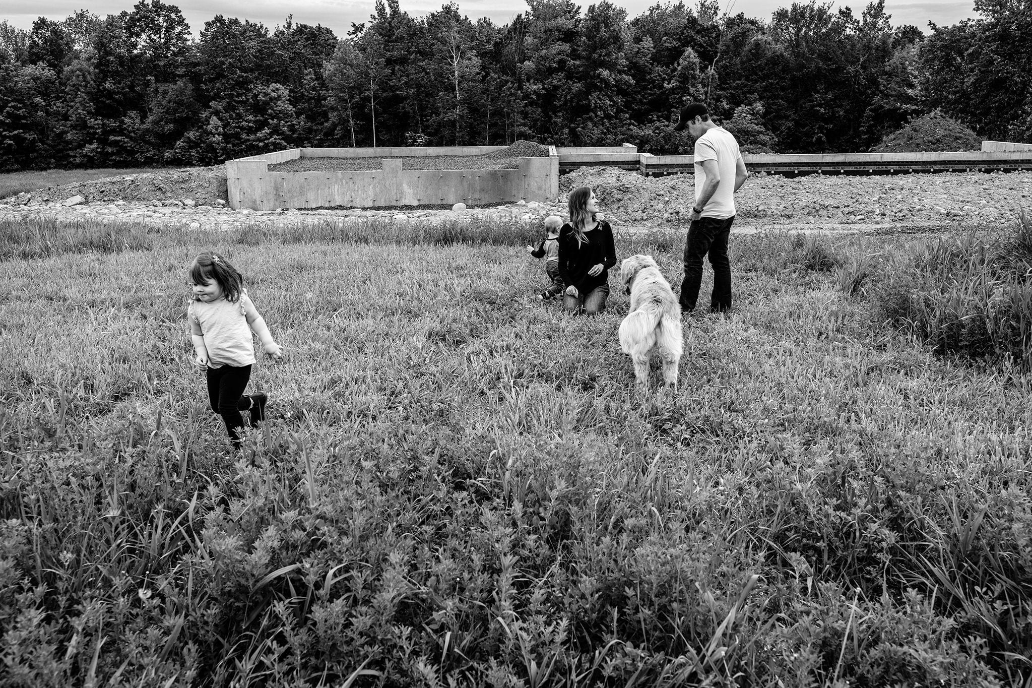 Cornwall milestone family photo at building site with girl running away from family in field while mom and dad sit with brother and dog