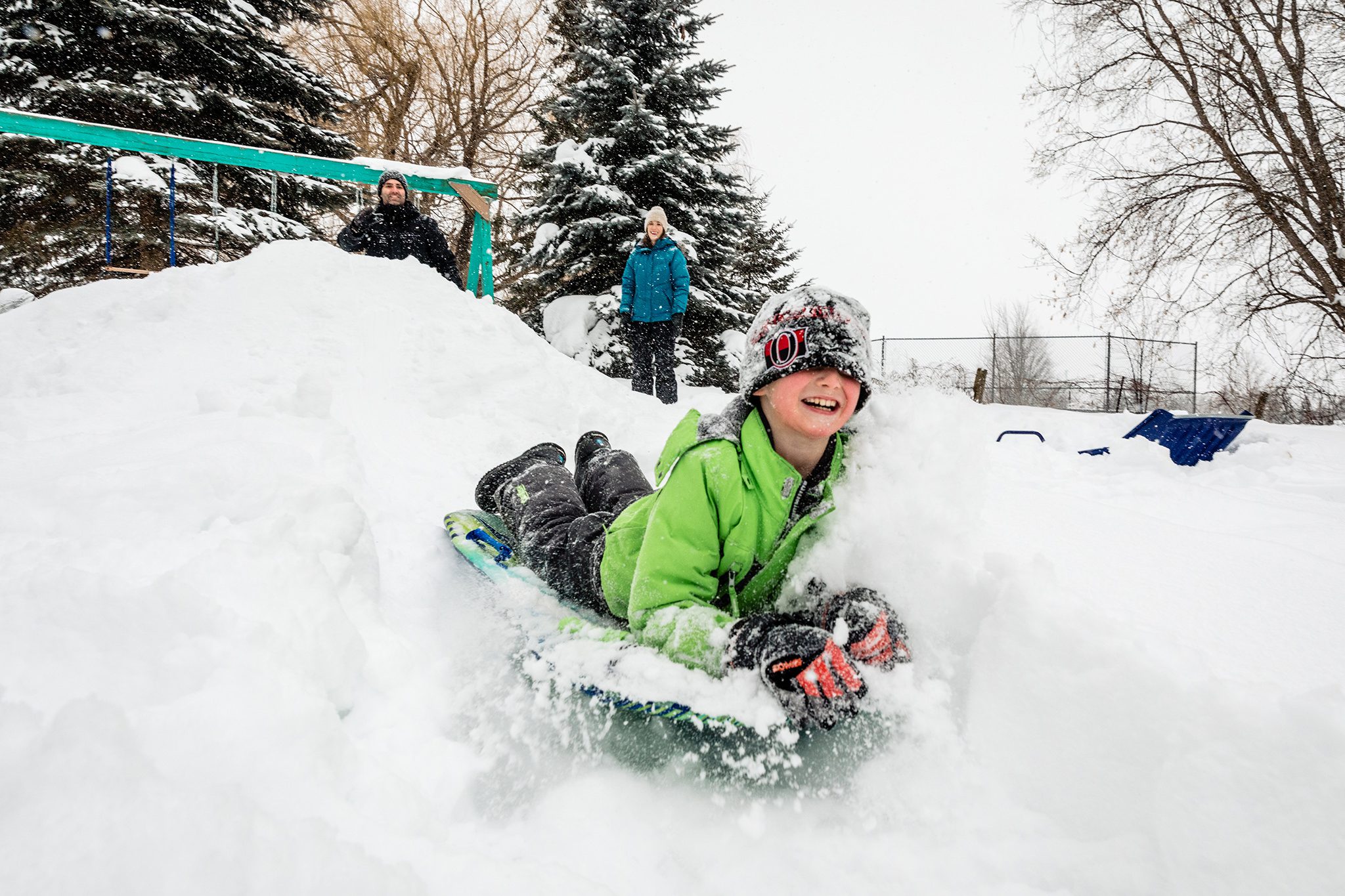 boy in green snow jacket slides down snowbank while hat covers eyes during winter documentary family photo session