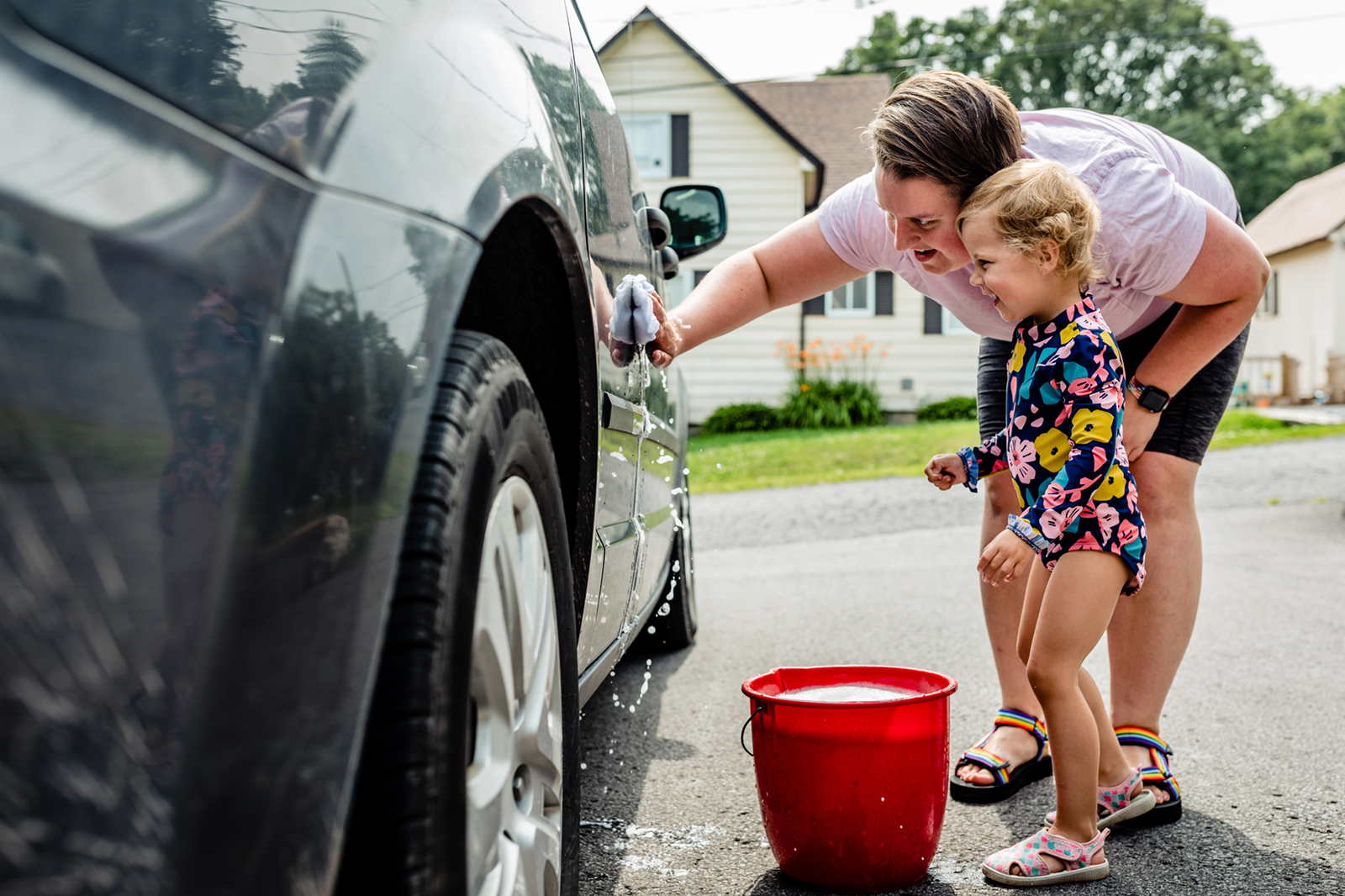 Woman and toddler girl laugh while washing black van with a soapy rag.