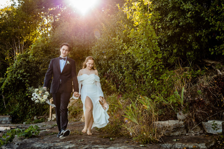 Bride holding her pair of heels and groom holding a bouquet of white flowers as they hold hands and walk beside a forest.