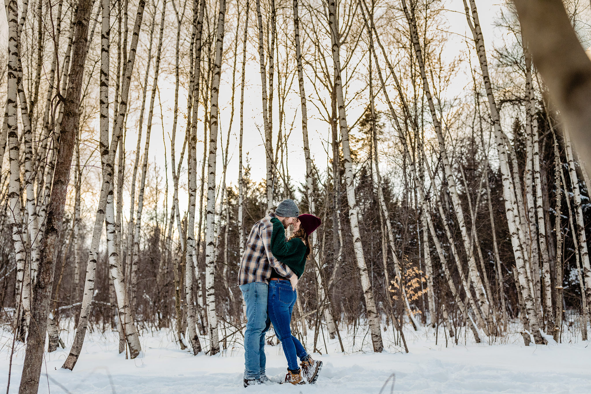 Newly engaged couple smiling and hugging surrounded by trees in a snowy forest.